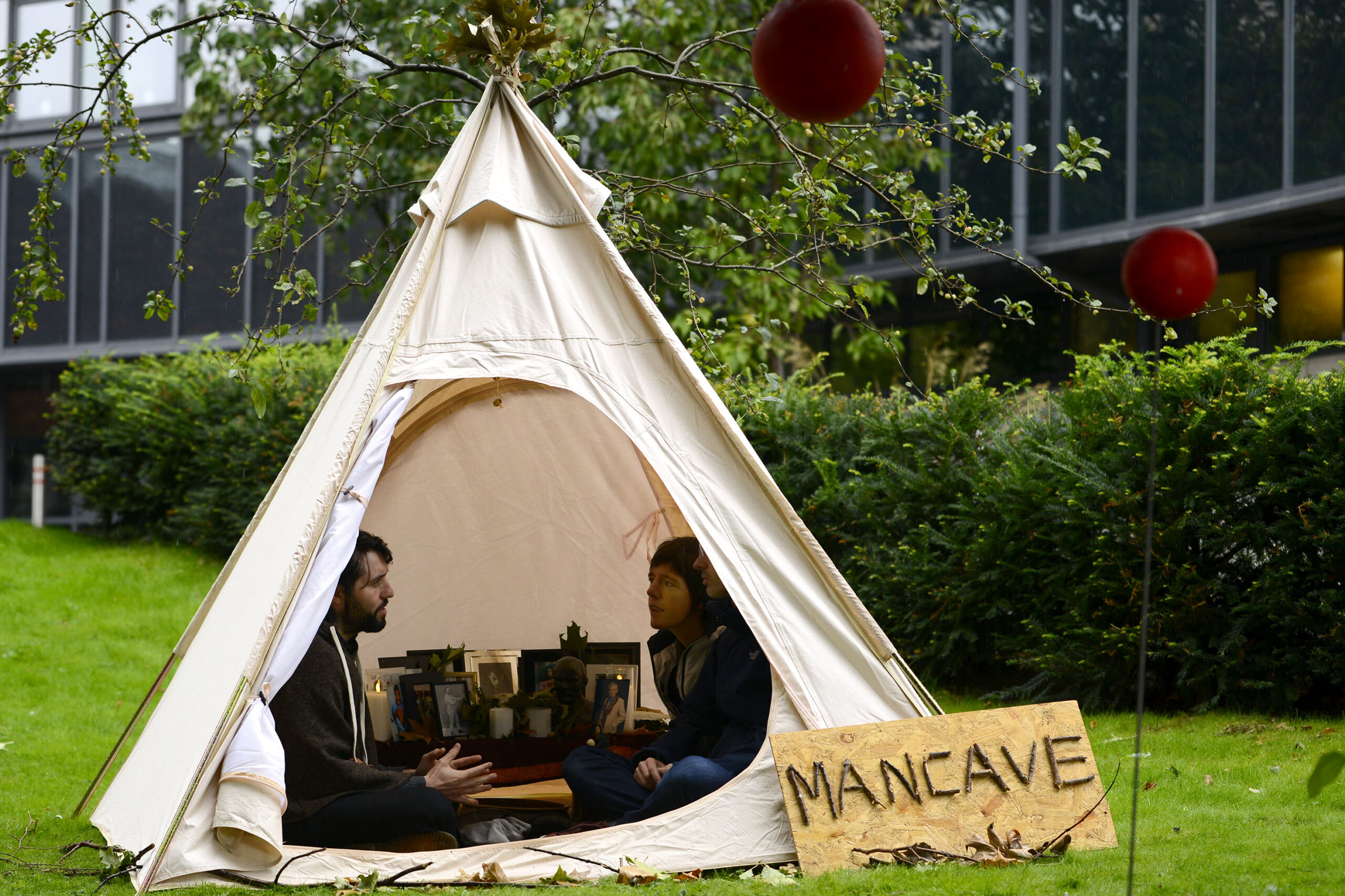 3 people talk in a small tent. Outside the tent there is a handmade sign on which is written "MANCAVE"